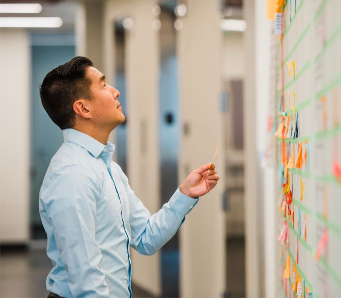 Man of Asian descent looking at sticky notes on an office whiteboard